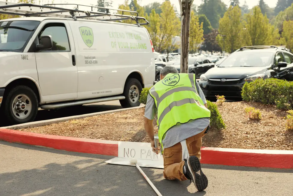 Technician in an HOA Services hi-vis vest stencilling NO PARKING on a red curb beside the lettered work van