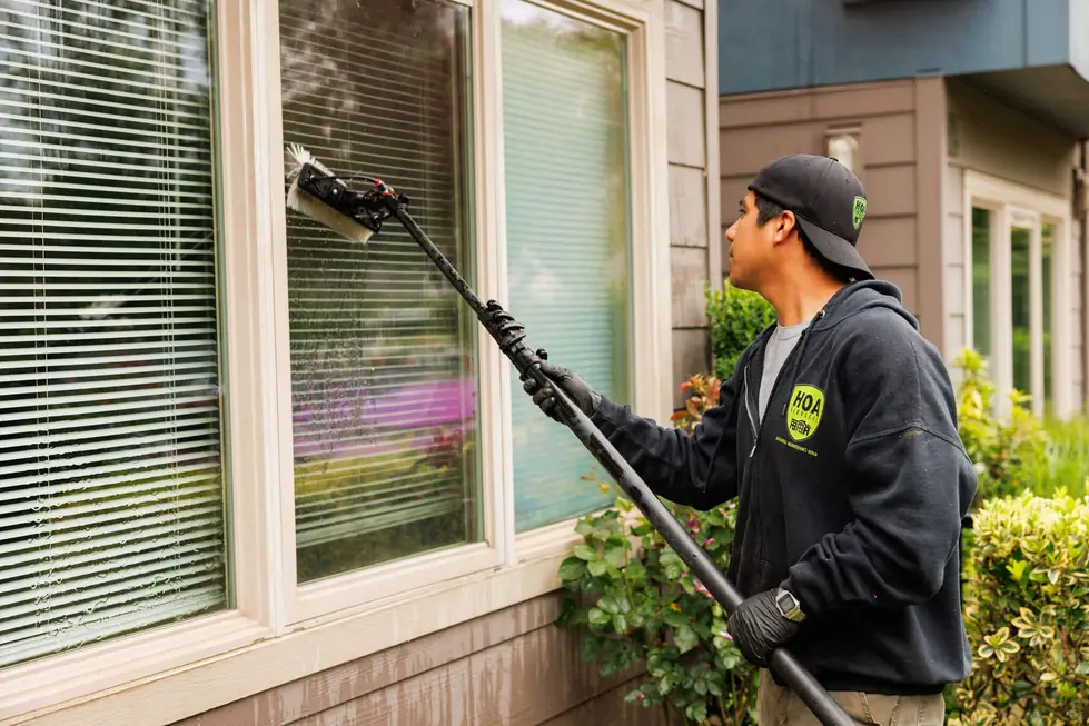 HOA Services technician in a branded hoodie pole-washing a ground-floor window beside planting beds